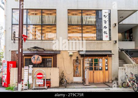 Two story Japanese restaurant in a white brick building. Entrance, name board and chochin, lantern, on the upper floor, unusual wooden window blinds. Stock Photo