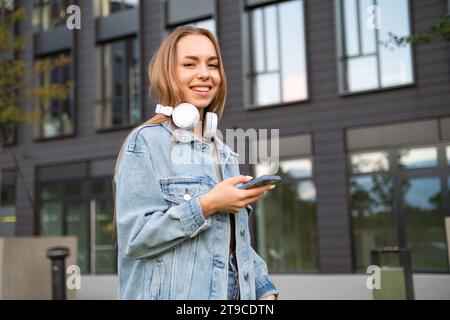 A grinning young woman, with her phone in hand and headphones stylishly draped around her neck, texting to her friend using smartphone  Stock Photo