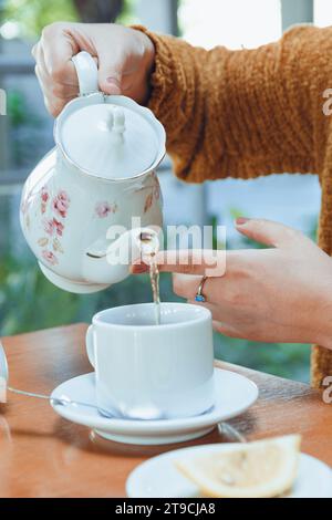 close-up of hands of unrecognizable Caucasian woman with white porcelain teapot pouring hot tea into white porcelain cup, on restaurant table Stock Photo