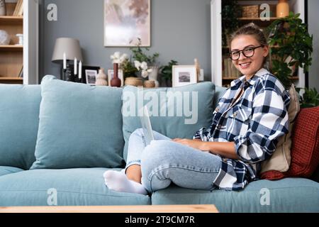 Young female freelancer in casual clothes sitting on comfortable couch and working remotely on laptop. Woman copyright writer or web developer work at Stock Photo