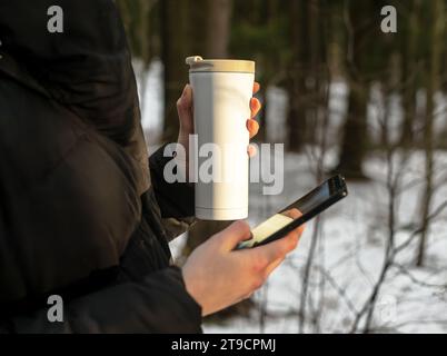 Hand holding thermo mug with hot tea, using mobile phone on winter holiday Stock Photo