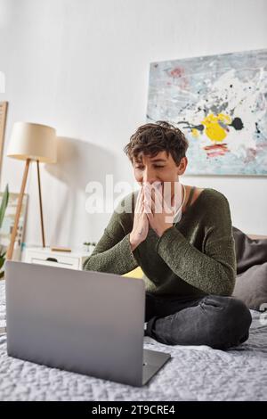 happy curly transgender blogger using laptop and sitting on bed in modern apartment, social media Stock Photo