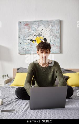 curly transgender blogger using laptop and sitting on bed in modern apartment, social media Stock Photo