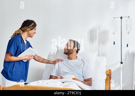 jolly african american nurse smiling happily at her indian patient that lying in hospital bed Stock Photo