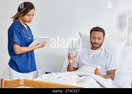 young african american nurse looking at tablet next to her indian patient holding pills, healthcare Stock Photo