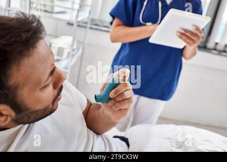 cropped view of indian man using asthma inhaler with blurred nurse on backdrop, healthcare Stock Photo