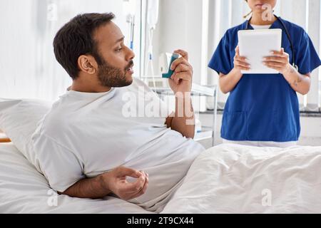 cropped view of indian man using asthma inhaler with blurred nurse on backdrop, healthcare Stock Photo
