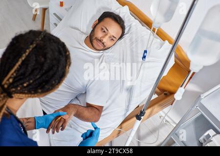 focus on indian man lying in hospital bed while african american nurse setting his drop counter Stock Photo