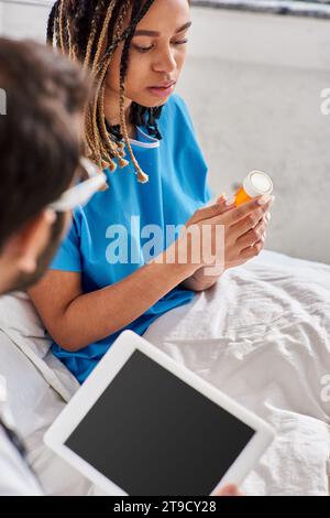 focus on ill african american woman sitting on bed and looking at her pills next to indian doctor Stock Photo