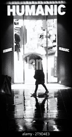 Black and White Art photo of a silhouette of a woman holding an umbrella passing in front of the Humanic store, Vienna, Austria during the rain Stock Photo