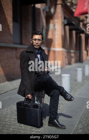 Office worker wearing black suit and carrying black briefcase sitting waiting for taxi on the roadside in the Old City of The Bund, Shanghai, China Stock Photo