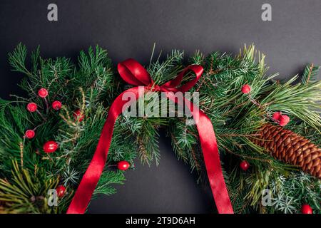 Christmas and New Year garland made of fir, pine evergreen branches, rose hips, cones decorated with red ribbon bow on black background. Festive decor Stock Photo