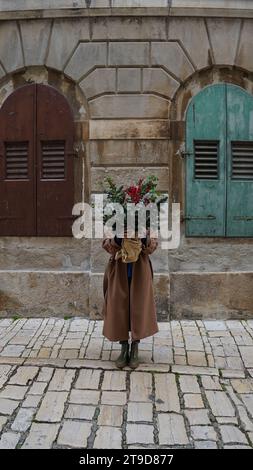 A girl in a beige coat holds a Christmas bouquet with branches of fir, winter berries, eucalyptus on the background of a stone house Stock Photo