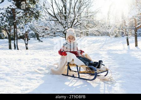Cute toddler boy sitting on the sleigh in a snowy city park during sunny winter day Stock Photo