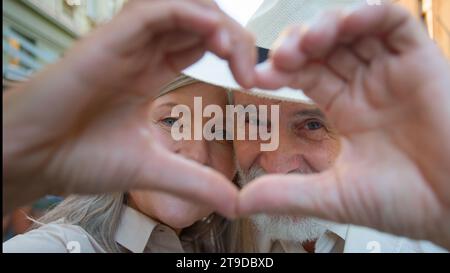 Close up portrait happy Caucasian woman man old retired family couple looking at camera through joined fingers heart gesture love feelings celebrate Stock Photo