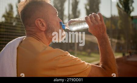 Back view old male sportsman athlete walking stadium outdoors drinking cold water from sports bottle Caucasian man pensioner drink quench thirst Stock Photo