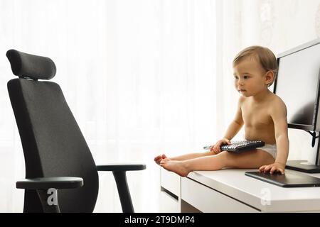 Little boy is sitting on a desk and playing with a keyboard in a home office workplace. Stock Photo