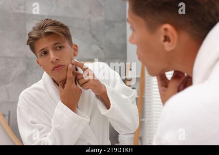 Upset young man looking at mirror and popping pimple on his face indoors. Acne problem Stock Photo