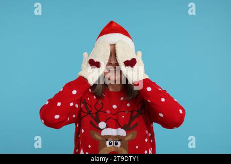 Senior woman in Christmas sweater and Santa hat covering face with hands in knitted mittens on light blue background Stock Photo