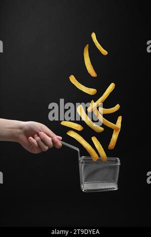 French fries falling into metal basket held by woman on black background, closeup Stock Photo