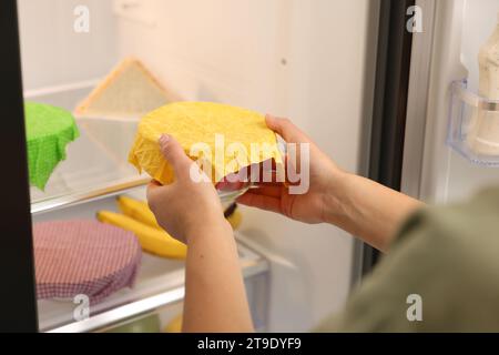 Woman putting bowl covered with beeswax food wrap into refrigerator, closeup Stock Photo