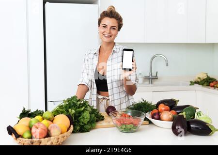 Young pretty woman is holding smartphone with blank screen for your design during cooking in modern kitchen Stock Photo