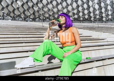 Carefree young woman wearing colorful sportswear is drinking water after her summer street workout Stock Photo