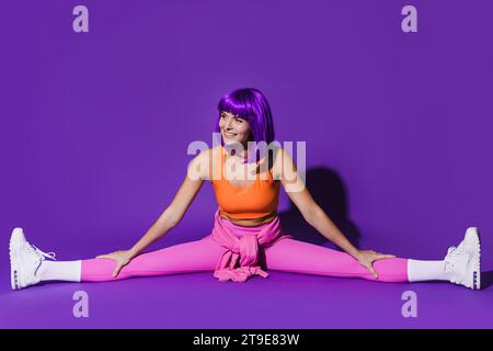 Young cheerful woman wearing colorful sportswear doing splits against purple background Stock Photo