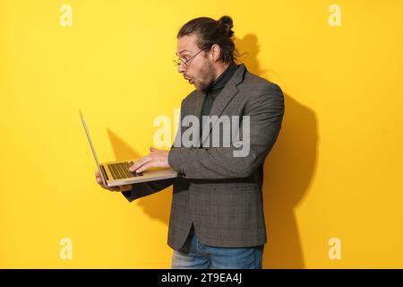 Surprised bearded man wearing eyeglasses is using laptop computer on yellow background Stock Photo