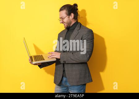 Handsome happy bearded man wearing eyeglasses is using laptop computer on yellow background Stock Photo