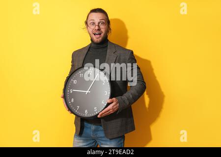Surprised bearded man wearing glasses holding big clock on yellow background Stock Photo