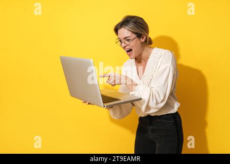 Cheerful young woman wearing eyeglasses is using laptop computer on yellow background Stock Photo