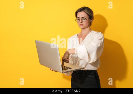 Young nerdy woman wearing eyeglasses is using laptop computer on yellow background Stock Photo