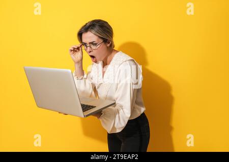Young suprised woman wearing eyeglasses using laptop computer on yellow background Stock Photo