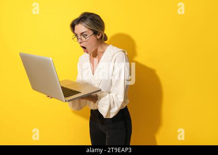 Young suprised woman wearing eyeglasses using laptop computer on yellow background Stock Photo