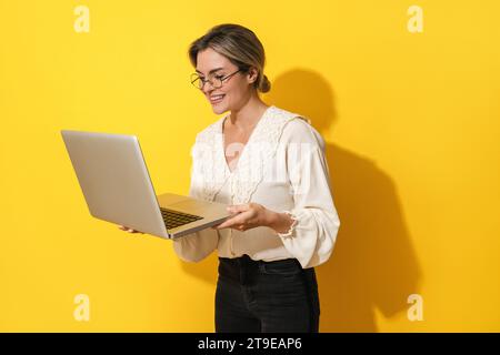 Cheerful young woman wearing eyeglasses is using laptop computer on yellow background Stock Photo