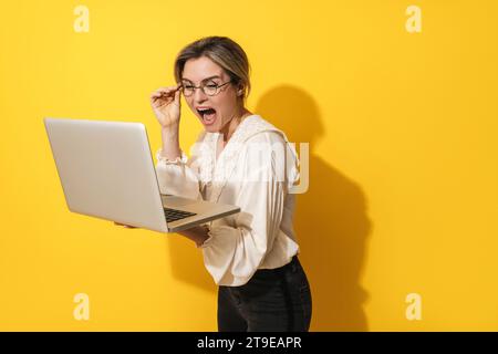 Cheerful young woman wearing eyeglasses is using laptop computer on yellow background Stock Photo