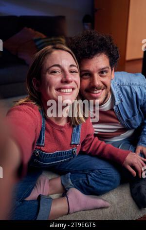 Young couple at home using smartphone taking selfie looking at camera smiling. Stock Photo