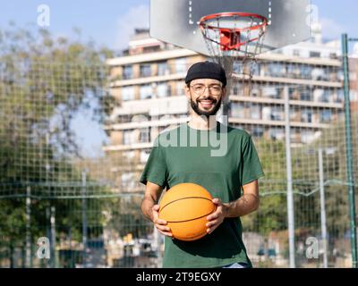 Portrait of a young hipster man with glasses smiling and holding a basketball in his hands looking at camera smiling on an urban basketball court Stock Photo
