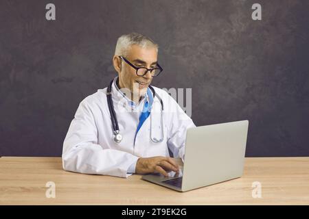 Portrait of smiling friendly senior male doctor providing online services using laptop. Stock Photo