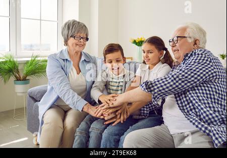 Grandparents sitting with grandchildren on sofa at home and putting their arms together in stack. Stock Photo