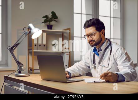 Male doctor sitting at desk, using laptop, studying information, and taking notes Stock Photo