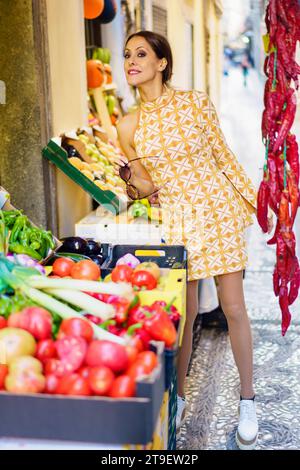 Full body of stylish young female model in casual trendy short cloth standing with assorted vegetables and fruits while posing with grocery in stall Stock Photo