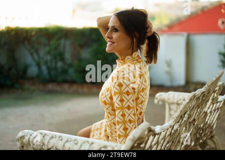 Side view of cheerful young female in stylish short gown sitting on wicker bench and holding hair on head, while looking away and resting in park on s Stock Photo