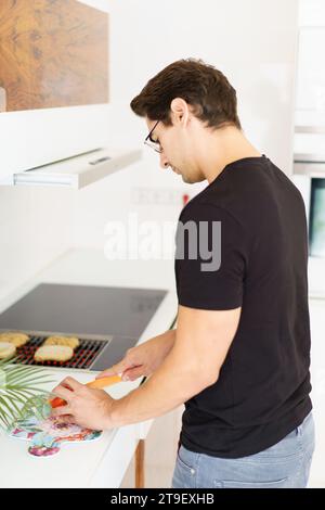 Side view of adult male in casual clothes and eyeglasses looking down while standing in kitchen near cooking range, with bread slices on grill and cut Stock Photo
