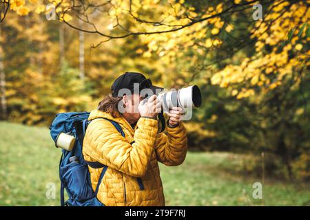 Hiker with backpack is wearing yellow jacket and photographing at his camera. Wildlife photographer in autumn forest Stock Photo