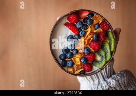 Woman holding healthy breakfast with fruits in coconut bowl. Yogurt with corn flakes and blueberry, strawberry, kiwi and raspberry. Vegetarian food Stock Photo