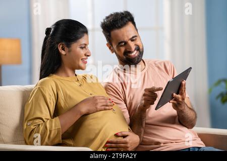 Happy Indian husband with pregnant wife discussing from digital tablet at home - concept of pregnancy preparations, togetherness and spending time Stock Photo