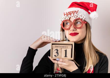 Beautiful, young, blonde girl with Santa hat and prop glasses holding a wooden calendar date box. December 31st Stock Photo