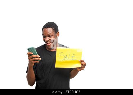 Young man holding yellow sale advertising poster board while using smartphone. Excited african man on black shirt with surprise face. Black Friday sal Stock Photo